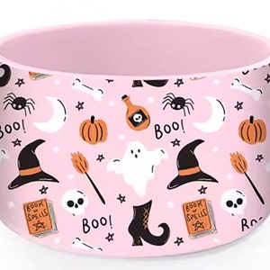 May include: Pink ceramic mug with a Halloween themed design. The mug features a repeating pattern of ghosts, witches hats, pumpkins, stars, moons, bones, and the word "Boo!"