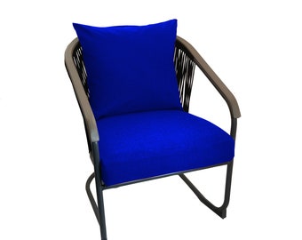 Könnte beinhalten: Moderner Gartenstuhl mit dunklem Metallrahmen und geschwungenen Armlehnen. Sitz und Rückenkissen sind in leuchtendem Blau gehalten, ergänzt durch ein passendes quadratisches Kissen. Der Stuhl hat geflochtene Seildetails auf der Rückseite.