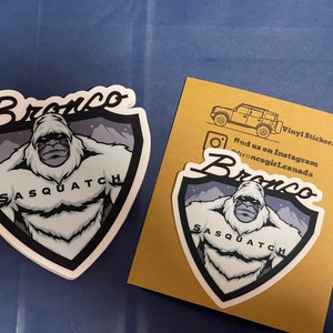 May include: Two white vinyl stickers with a black outline featuring a cartoon Sasquatch with the text "Bronco Sasquatch" in a white, black, and gray color scheme. The stickers are on a brown background. Find us on Instagram @broncogirl.canada.