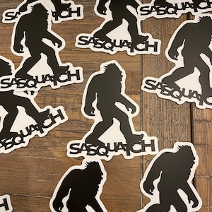 Peut inclure: Autocollants noirs et blancs représentant une silhouette de Sasquatch en mouvement. Le mot "SASQUATCH" est imprimé en blanc sous chaque figure. Les autocollants sont disposés sur une surface en bois.