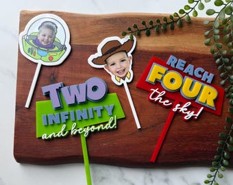 Puede incluir: Un conjunto de tres adornos personalizados para pasteles que incluyen un casco de Buzz Lightyear con la cara de un niño, un sombrero de Woody con la cara de un niño, un letrero rojo con el texto REACH FOUR the sky y un letrero verde con el texto TWO INFINITY and beyond.