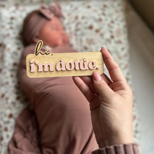 May include: A wooden sign with the text "hi, i'm dottie." in pink letters. The sign is being held by a hand.