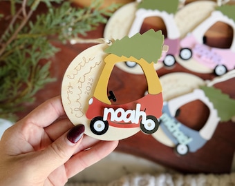 Op de afbeelding: Houten kerstversiering met een rode en gele auto met de naam "Noah" en een groene boom erop. De versiering staat op een houten schijf met de tekst "a cozy little Christmas 2025". Andere ornamenten zijn zichtbaar op de achtergrond.