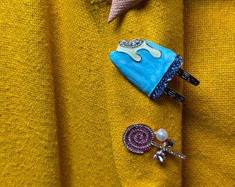 Puede incluir: Una chaqueta de tela amarilla con cuatro alfileres decorativos. Los alfileres tienen forma de cono de helado, paleta de hielo, piruleta y caramelo. Los alfileres están cubiertos de pedrería.