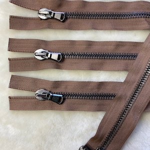 Puede incluir: Cuatro cierres de cremallera marrones con dientes metálicos y tiradores negros. Las cremalleras están dispuestas horizontalmente sobre una superficie blanca y peluda.