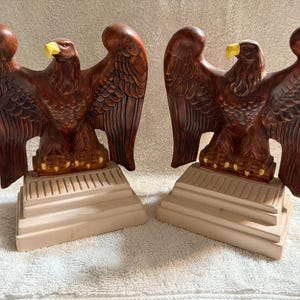 Op de afbeelding: Twee bruine keramische adelaarsbeelden met uitgespreide vleugels en gele snavels. Elke adelaar staat op een beige trapvormige basis. De beelden hebben gebeeldhouwde veerdetaillering.