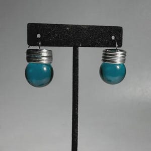 May include: A pair of teal glass ball earrings with silver-colored metal accents. The earrings are displayed on a black earring stand against a gray background. The glass balls are round and have a smooth surface.