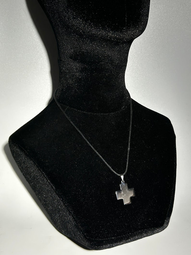 May include: A silver cross pendant necklace on a black cord, displayed on a black velvet mannequin bust.