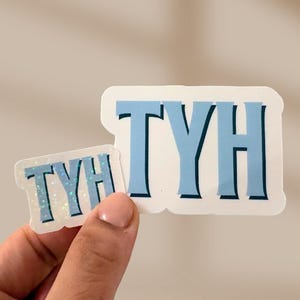 Può includere: Due adesivi con le lettere "TYH" in azzurro con un contorno blu scuro. L'adesivo più grande è tenuto davanti a uno sfondo neutro. L'adesivo più piccolo è tenuto davanti a quello più grande.