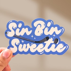 May include: A blue sticker with the words "Sin Bin Sweetie" in white, stylized bubble letters. A hockey stick and puck are featured in the design. The sticker is held by a hand.