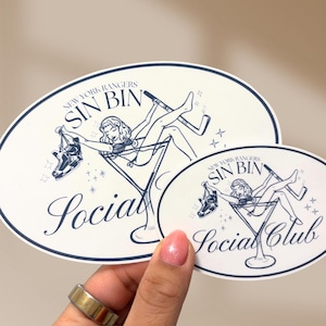 May include: Two oval stickers with a navy blue and white design. The stickers feature a woman in a martini glass with hockey skates and a hockey stick. The text reads "New York Rangers Sin Bin Social Club."