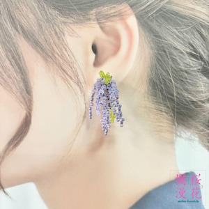 以下が含まれることがあります： 藤の花の形をしたビーズのイヤリング。イヤリングは、緑と黄色のアクセントが付いた小さな紫色のビーズが特徴です。イヤリングは人の耳の隣に表示されています。