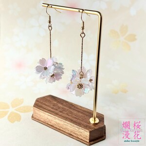以下が含まれることがあります： イリデセントな花型のビーズのクラスターが特徴のぶら下がりピアス。ピアスはゴールドカラーのフックとチェーンから吊り下げられています。ゴールドカラーの金属製サポートが付いた木製のスタンドに展示されています。