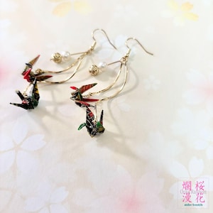 以下が含まれることがあります： 赤、緑、黒の模様が入った折り鶴のペンダントが付いた、ゴールドトーンのドロップピアス。ピアスには、繊細で芸術的なデザインを作り出す、小さなパールとゴールドビーズのアクセントも含まれています。