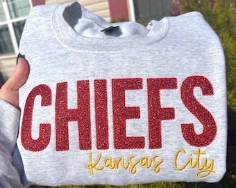 Peut inclure: Un sweat-shirt gris clair avec le mot "CHIEFS" en grandes lettres rouges scintillantes. En dessous, les mots "Kansas City" sont brodés en or. Le sweat-shirt est de style col rond.