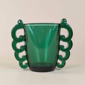 以下が含まれることがあります： 2つの装飾的なスクロール型のハンドルが付いた、濃い緑色のガラスの花瓶。花瓶は円筒形の本体と暗いベースを持っています。ハンドルは明るい緑色で、デザインに装飾的な要素を追加しています。花瓶は空です。