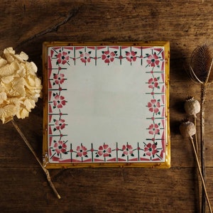 May include: A white ceramic trivet with a yellow border and a floral design in pink and green. The trivet is rectangular and has a raised edge.