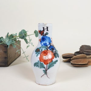 May include: A white ceramic pitcher with a floral design in shades of blue, orange, and green. The pitcher has a small spout and a handle. The letter "H" is painted on the front of the pitcher.