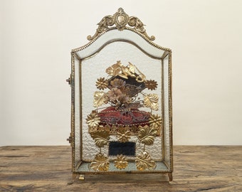 Op de afbeelding: Een glazen vitrine met een gouden frame en sierlijke details. In de vitrine bevindt zich een rood fluwelen kussen met een bloemstuk van gedroogde bloemen en gouden accenten.