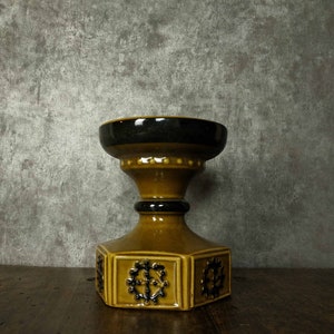 Könnte beinhalten: Ein brauner und schwarzer Kerzenhalter aus Keramik mit geometrischem Design. Der Halter hat eine breite Basis und eine schmale Oberseite mit einer runden Öffnung.