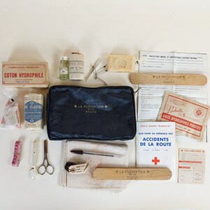 Op de afbeelding: Een vintage EHBO-doos met een blauwe canvas tas, gaas, katoen, verbanden en diverse andere medische benodigdheden. De doos bevat de tekst "La Prevention de la Route", wat vertaald wordt als "Verkeersveiligheid".