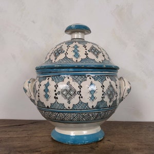 May include: A white ceramic serving dish with a blue lid and intricate geometric patterns. The dish has two handles and a blue base.