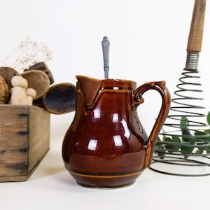 May include: A brown ceramic creamer with a handle and a spoon inside. The creamer has a glossy finish and a slight lip around the opening.
