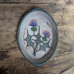 May include: An oval ceramic dish with a speckled off-white base and a dark teal rim. The center features a hand-painted illustration of two purple thistle flowers with green spiky leaves and stems, accented by brown splatters on the surface.