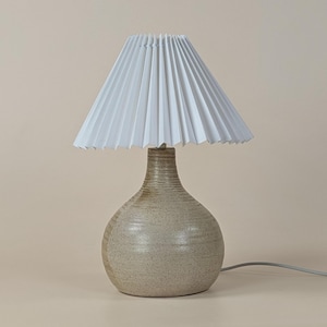 Op de afbeelding: Een keramische tafellamp met een witte geplooide lampenkap. De lampvoet is een gespikkelde beige kleur met een afgeronde vorm en een textuur. Een grijze stroomkabel komt uit de basis.