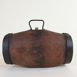 Peut inclure: Une boîte en cuir marron en forme de tonneau avec une poignée en métal et des bandes métalliques autour des extrémités. La boîte est fermée et a un aspect usé et vintage.