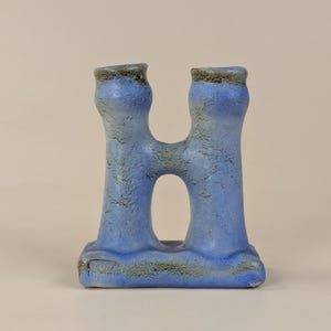 May include: A blue ceramic sculpture with a unique, open design. The piece features two vertical columns connected at the top and base, with a central opening. The surface has a textured appearance with darker accents, creating visual interest.