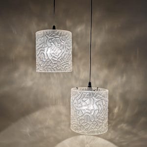 Puede incluir: Dos lámparas colgantes cilíndricas con pantallas de vidrio transparente texturizado y un intrincado patrón abstracto en relieve. Cada lámpara cuelga de un cable negro fino, emitiendo un brillo suave que proyecta sombras estampadas alrededor.