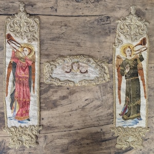 May include: Three embroidered fabric panels featuring gold thread detailing. Two vertical rectangular panels depict standing angels playing trumpets, one in a red robe and one in a green robe. A central horizontal panel shows three cherub faces.