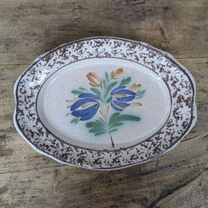 Puede incluir: Una bandeja ovalada de cerámica con una base de color crema y un acabado de esmalte craquelado fino. El borde está decorado con un patrón moteado de color marrón oscuro, mientras que el centro muestra un motivo floral pintado a mano.