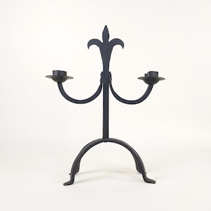 May include: A black wrought iron candle holder with two arms and a fleur-de-lis design. The candle holder has a sturdy base with three feet and is designed to hold two candles. The overall design is gothic and decorative.