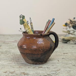 Può includere: Un vaso di ceramica marrone con manico, pieno di matite, un righello e fiori secchi. Il vaso ha una superficie strutturata con fasce marrone scuro e nere. Gli oggetti all'interno sono visibili.