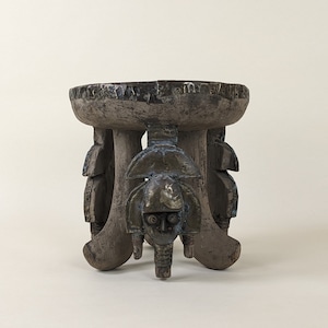 May include: A wooden stool with a carved design. The stool has four legs and a circular top. The top is decorated with a carved design of a human face.