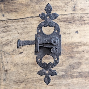May include: A black, antique-style metal latch with decorative cutouts and a handle. The latch is mounted on a wooden surface, showcasing its rustic design and aged appearance. The latch measures approximately 6 inches in length.