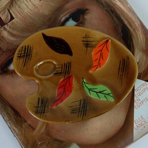 Può includere: Una tavolozza da artista in ceramica marrone con un foro per il pollice e linee nere. La tavolozza è decorata con quattro foglie dipinte in tonalità di rosso, arancione, verde e marrone.