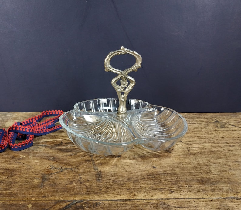 May include: A clear glass serving dish with three sections and a silver handle. The dish is shaped like a clover leaf and has a decorative design.