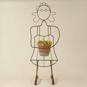 May include: A metal plant stand shaped like a person with glasses and a flower pot in their hands. The stand is made of thin metal and has a rustic brown finish.