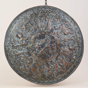May include: A large, round, antique metal shield with a detailed relief depicting a battle scene. The shield features a central design surrounded by intricate patterns and figures in shades of bronze and blue-gray.