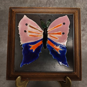 Peut inclure: Un carreau de céramique encadré avec un motif de papillon coloré. Le papillon a des ailes roses avec des accents orange et noirs, et des détails bleus et noirs sur le corps. Le carreau est encadré dans un cadre en bois foncé.