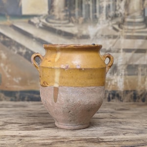 Op de afbeelding: Een aardewerken pot met twee oren, voorzien van een geglazuurde mosterdgele bovenkant die uitloopt over een textuurrijke, ongeglazuurde terracotta basis. De pot heeft een brede, uitlopende rand en een ronde vorm, met een duidelijk contrast tussen de gladde, glanzende bovenkant en de ruwe, matte onderkant.