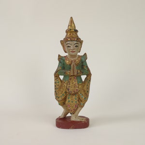 Puede incluir: Una figurita de madera de una persona con un traje tradicional tailandés. La figurita está pintada en oro, verde y rojo, y tiene una corona en la cabeza. La persona está de pie con las manos juntas frente a ella.