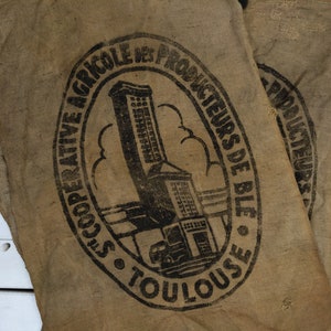 Puede incluir: Un saco de arpillera marrón con un sello negro que dice "Coopérative Agricole des Producteurs de Blé de Toulouse". El sello presenta un edificio con una torre alta y una nube.