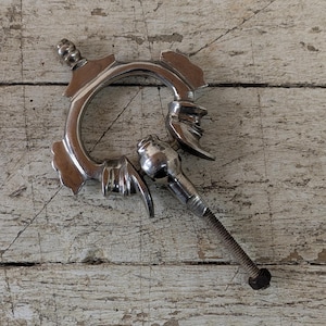 Puede incluir: Un llamador de puerta de metal plateado con un diseño decorativo que presenta una cara y un asa de anillo. El llamador está unido a un tornillo.