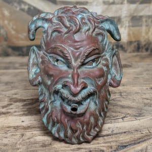 May include: A bronze-toned metal wall fountain sculpture depicting a horned, bearded face with a weathered patina of green oxidation. The figure features pointed ears, a wide grin, and a small circular spout protruding from the center of the mouth.