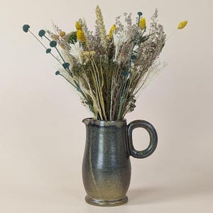 May include: A bouquet of dried flowers in shades of yellow, white, and blue arranged in a dark green ceramic pitcher.