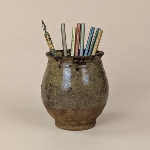Può includere: Un vaso in ceramica rustico, fatto a mano, con una glassa screziata verde e marrone strutturata. Il vaso contiene diverse matite e una penna decorativa. Le matite sono di vari colori e la penna ha un motivo.